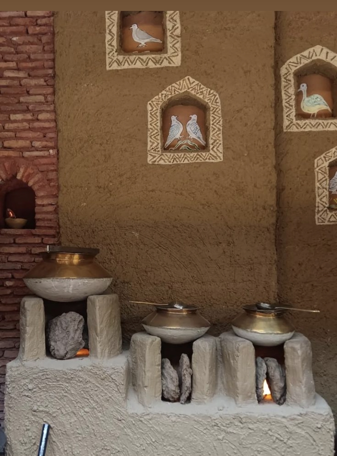 Mud wall with hand-painted dove niches above brass pot chulha stations